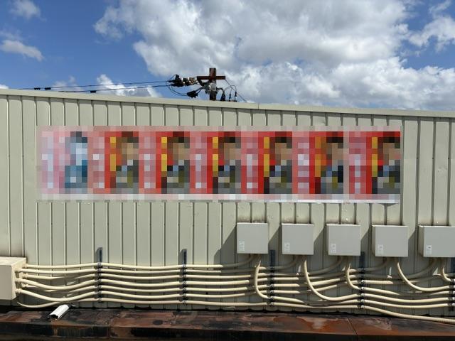 Row of blurred portraits displayed on a beige metal wall, with utility boxes and cables below under a blue sky.